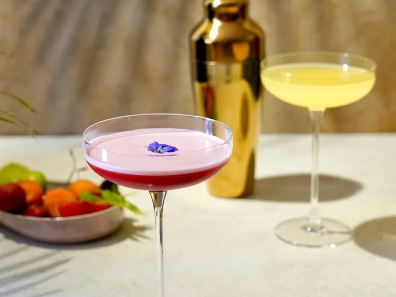 Imagen de referencia: cu{00F3}ctel rosa en copa coupe con una flor comestible, junto a una coctelera dorada